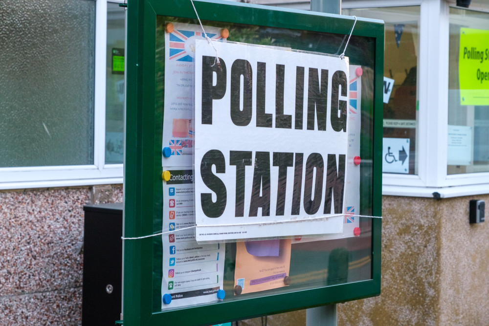 A black and white polling station sign affixed to a noticeboard in front of the village hall.
