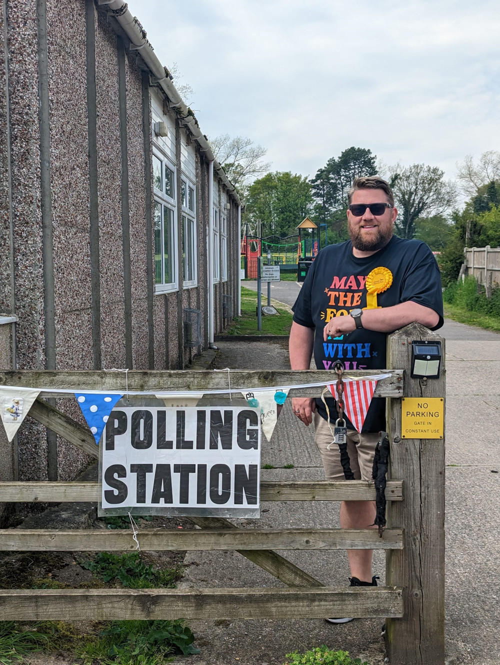 Lib Dem candidate Lee Castle wearing a rosette, standing next to polling station sign outside the village hall.